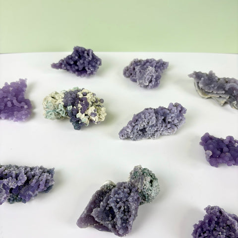 Collection of raw grape agate clusters featuring deep purple, botryoidal crystal formations on a white surface.