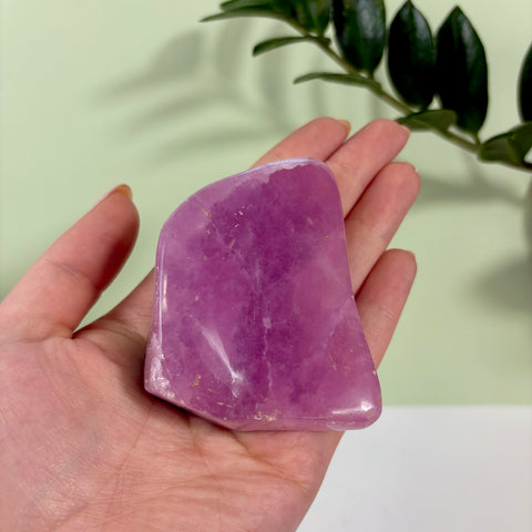 Polished jelly pink aragonite free form crystal held in a hand against a light green background.