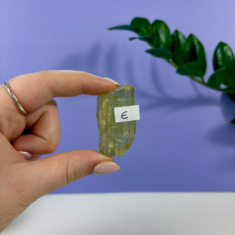 Hand holding a raw, translucent lime kunzite crystal with a label marked E against a purple background.
