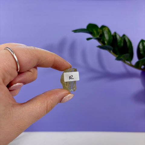 Hand holding a raw, translucent lime kunzite crystal labeled R against a purple background with a green plant.
