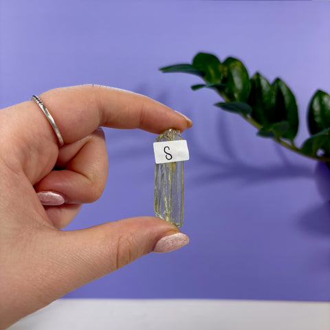 Small, translucent lime kunzite crystal held between fingers against a purple background with a size S label.