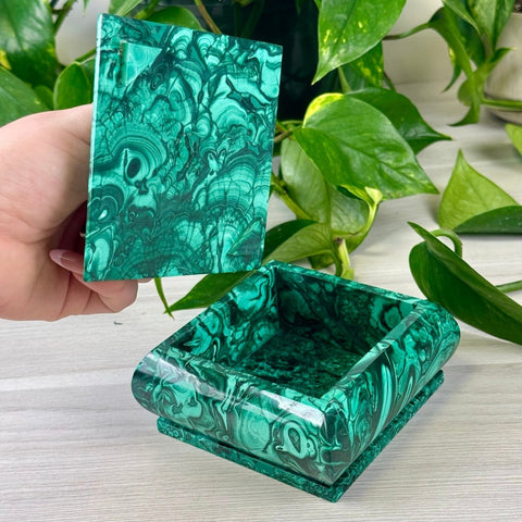 Square malachite jewellery box with a vibrant green swirled pattern and a removable lid held by a hand.