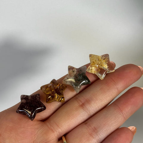 Four mini garden quartz star crystals, varying from dark brown to clear with golden inclusions, held in a hand.