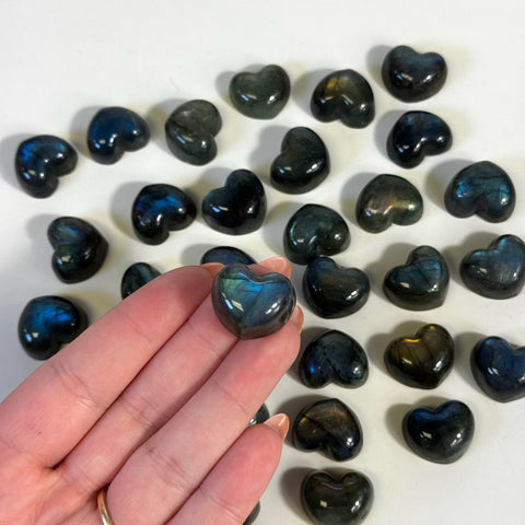 Hand holding a small, polished labradorite heart with blue and green flash, surrounded by many similar mini labradorite hearts.