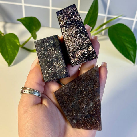 Three dark, glittery Mystery Galaxy Iolite crystals, two rectangular and one triangular, held in a hand.