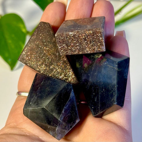 Hand holding four faceted Mystery Galaxy Iolite crystals: two dark blue, one sparkling brown cube, one sparkling brown triangle.