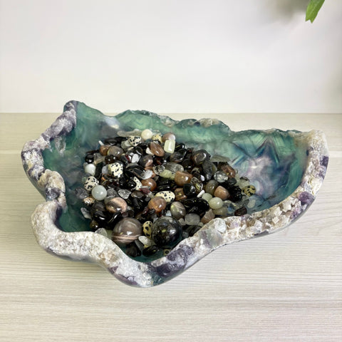 Large, organic-shaped fluorite bowl featuring purple and green hues filled with a variety of polished black and speckled stones.