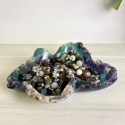 Polished fluorite bowl with purple and teal hues, filled with a collection of assorted smooth gemstone pebbles.