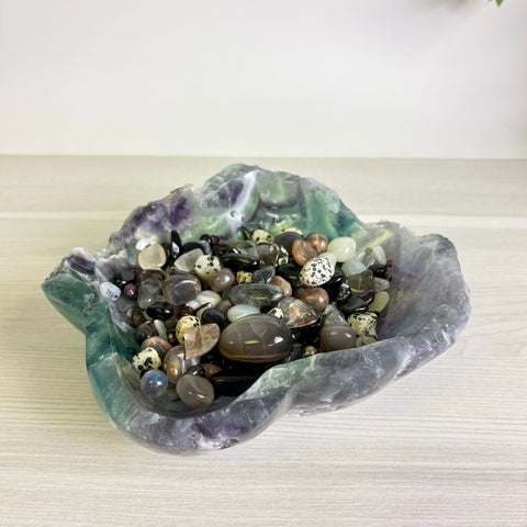 Polished fluorite crystal bowl filled with a variety of smooth, multi-colored tumbled gemstones on a wooden surface.