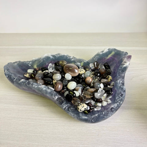 Hand-carved purple fluorite bowl filled with a variety of polished, multi-colored gemstone tumbles and crystals.