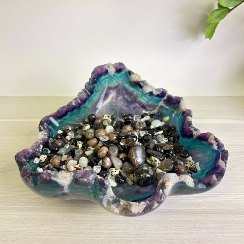 Polished fluorite crystal bowl in purple and teal hues filled with a variety of small, smooth, multi-colored gemstone pebbles.