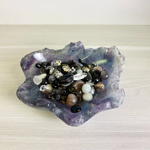 Hand-carved purple and green fluorite crystal bowl filled with a variety of polished black, white, and spotted gemstones.