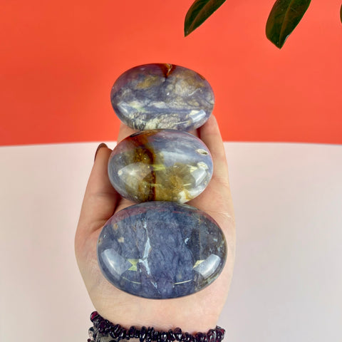 Three polished Nightshade Chalcedony palm stones with purple and golden inclusions held in a hand.