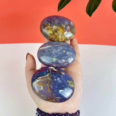 Three polished, oval Nightshade Chalcedony palm stones with deep purple, blue, and golden-brown marbled patterns held in a hand.