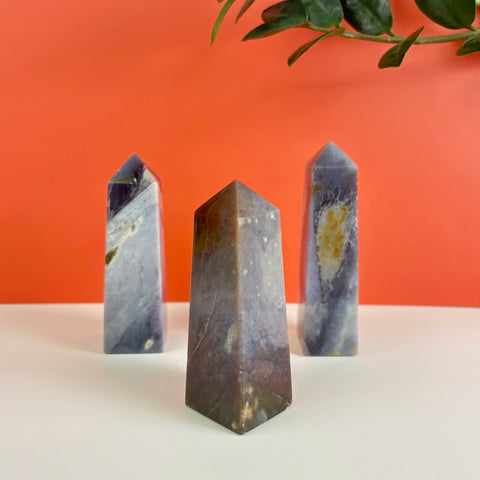 Three polished Nightshade Chalcedony crystal towers with purple and grey hues against a vibrant orange background.