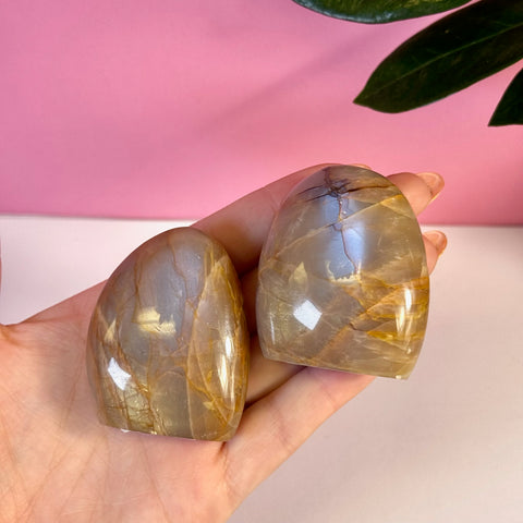 Two peach moonstone free forms with brown veins, held in a hand against a pink background.