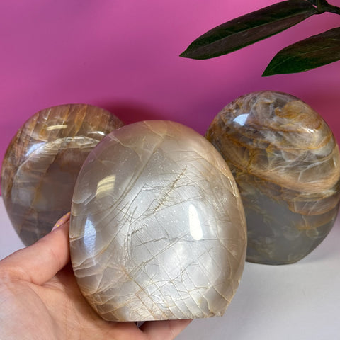 Polished peach moonstone free form with iridescent sheen, held in hand, with two similar stones in background.
