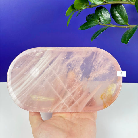 Oval rose quartz crystal tray with natural translucent pink veining held in a hand against a blue background.