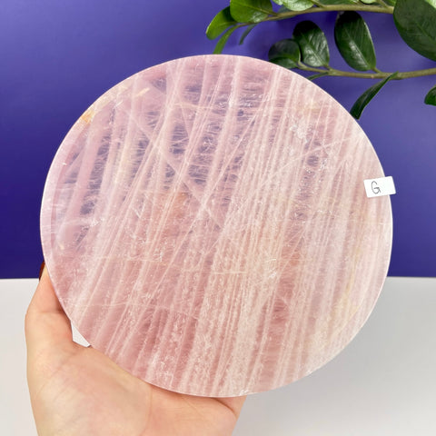 Round rose quartz plate with natural pink crystalline patterns held in a hand against a purple background.