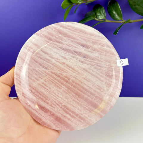 Round rose quartz crystal plate with natural pink veining held in a hand against a purple background.