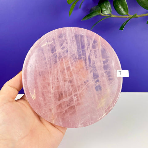 Round rose quartz crystal plate with natural veining held in a hand against a blue and white background.