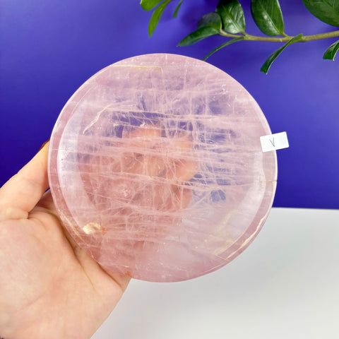 Hand holding a round, polished rose quartz crystal plate with natural pink veining against a purple background.