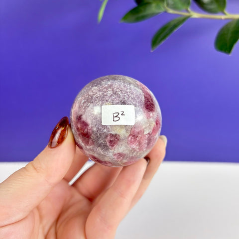 Polished pink tourmaline sphere with natural mineral inclusions held in hand against a purple background.
