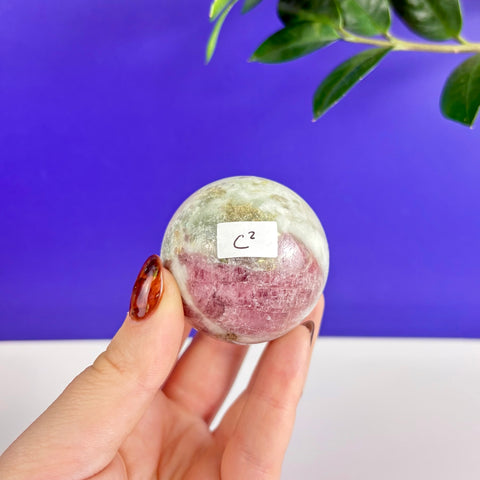 Polished pink and green tourmaline crystal sphere held in hand against a blue background.