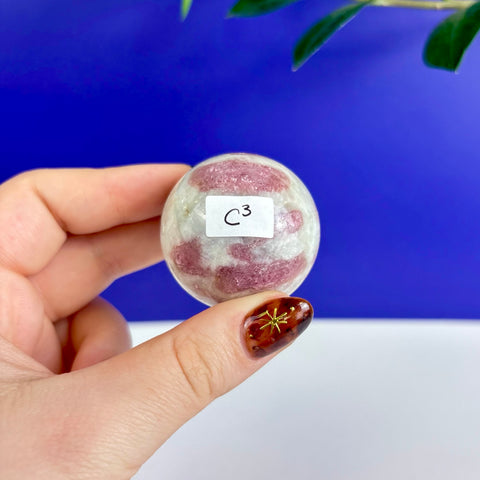 Polished pink tourmaline sphere with white quartz matrix held in hand against a bright blue background.