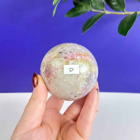 Polished pink tourmaline sphere with white and grey mineral inclusions held in a hand against a blue background.