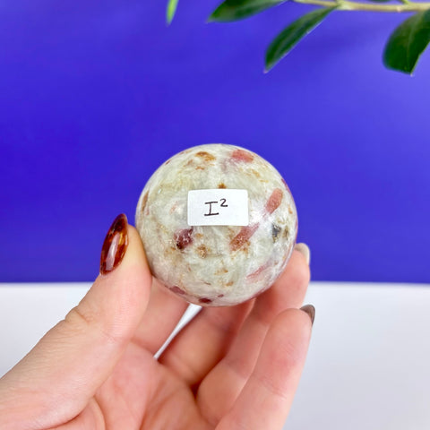Polished pink tourmaline sphere with white and grey inclusions held in a hand against a blue background.