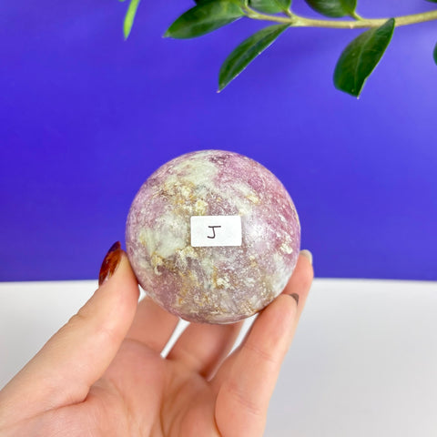 Polished pink tourmaline sphere with white mineral inclusions held in a hand against a blue and white background.