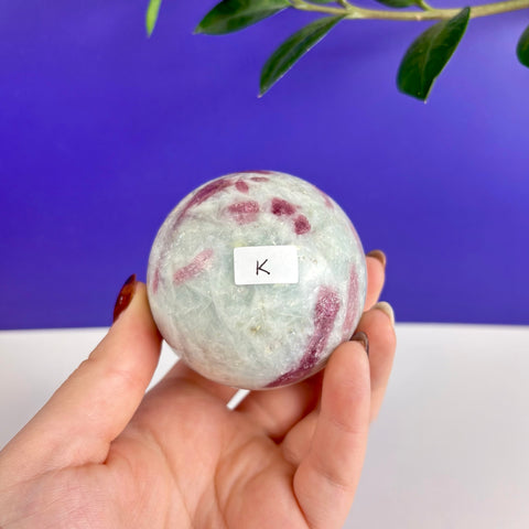 Polished pink tourmaline sphere with vibrant magenta inclusions held in a hand against a purple background.