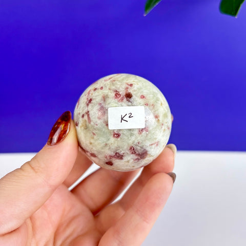 Polished pink tourmaline sphere with green mineral inclusions held in a hand against a blue and white background.