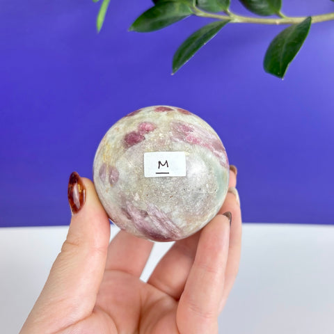 Polished pink tourmaline sphere with natural inclusions held in a hand against a blue and white background.