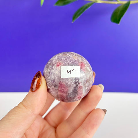 Polished pink tourmaline crystal sphere with natural inclusions held in a hand against a blue background.