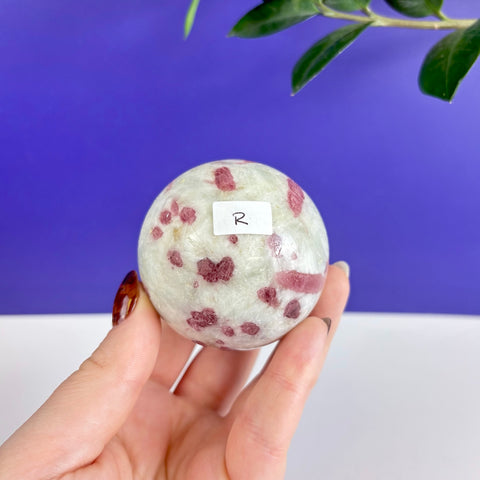 Polished pink tourmaline sphere with deep magenta inclusions held in a hand against a blue and white background.