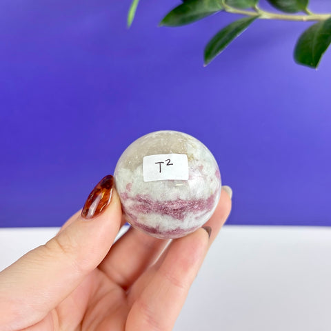 Polished pink tourmaline sphere with white mineral inclusions held in a hand against a blue background.