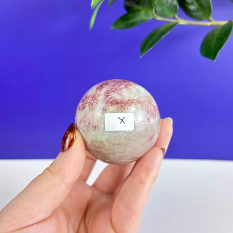 Polished pink tourmaline sphere with white mineral inclusions held in a hand against a blue and white background.