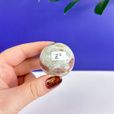 Polished pink tourmaline sphere with white and grey mineral inclusions held in a hand against a blue background.