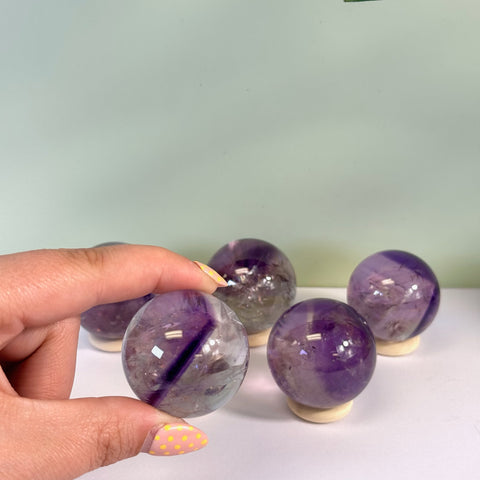 Hand holding a polished phantom amethyst sphere with purple and clear layers. Four more spheres sit on wooden stands.