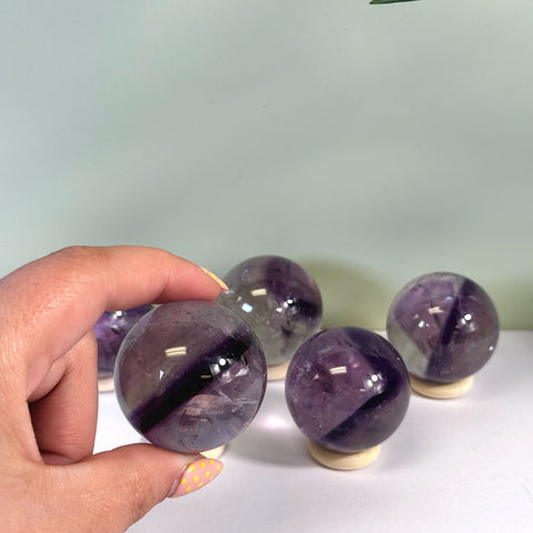 Hand holding a polished purple phantom amethyst sphere with white inclusions, four more spheres in background.