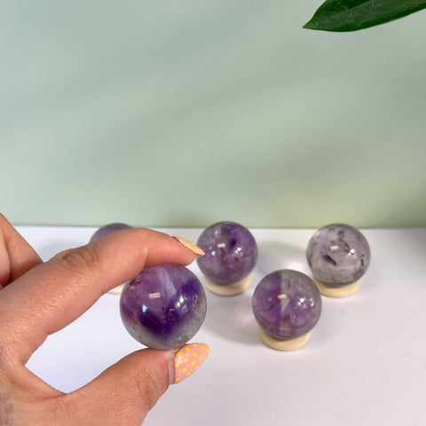 Hand holding a polished phantom amethyst sphere with deep purple inclusions, next to several others on small wooden stands.