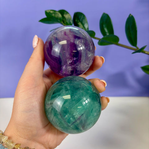 Hand holding a polished purple and green phantom fluorite sphere with layered mineral inclusions.