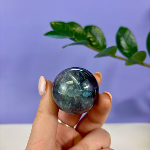 Hand holding a polished phantom fluorite sphere with deep blue and green layered mineral inclusions against a purple background.