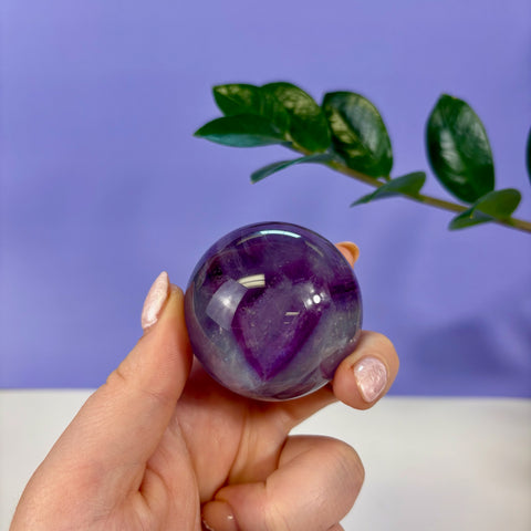 Hand holding a polished purple phantom fluorite sphere with visible internal growth layers against a purple background.