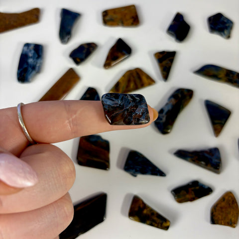 Polished blue and brown Pietersite slice held on a finger, surrounded by various loose gemstone pieces on a white background.