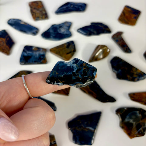 Polished blue and gold Pietersite slice held on a finger, surrounded by various loose gemstone pieces on a white surface.