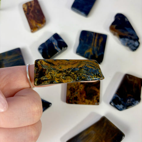 Polished rectangular Pietersite slice with swirling blue and golden-brown chatoyant patterns held by a finger.