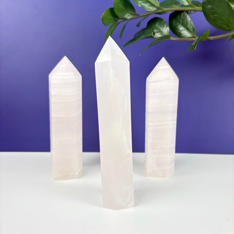 Three polished pink calcite crystal towers with banded textures standing on a white surface against a purple background.